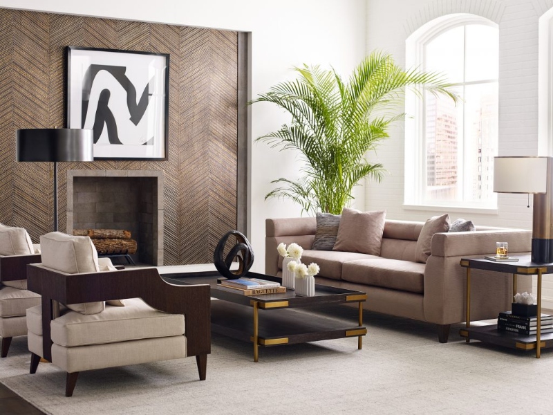 Rug Décor with Import Temptations. This modern contemporary living room has as a center piece a fireplace with a wall in wood, a brown light sofa, two armchairs in neutral with brown details, a black coffee table, and a neutral rug that covers all of the room.