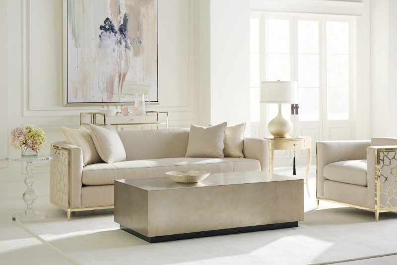 Rug Décor with Import Temptations. This white modern living room has a simple and neutral rug, a neutral sofa and armchair with gold details, a silver coffee table, and a painting in light colors.